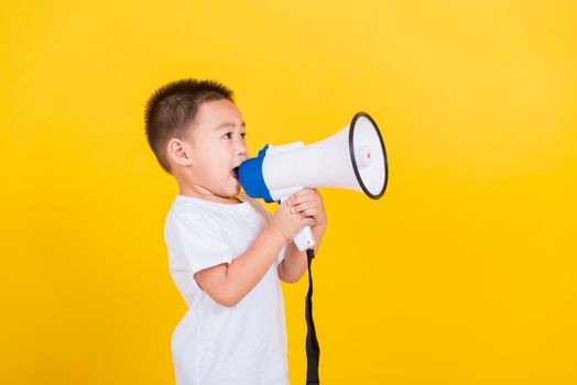 Asian Thai happy portrait cute little cheerful child boy holding and shouting or screaming through the megaphone her looking to side, studio shot isolated on yellow background with copy space