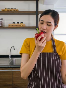 The housewife dressed in an apron bites the delicious red bell pepper. Morning atmosphere in a modern kitchen