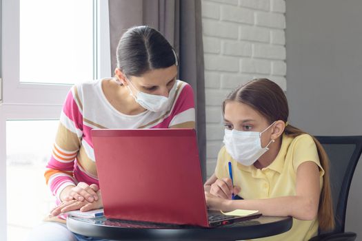 The tutor at home in a medical mask explains to the girl the course of solving homework