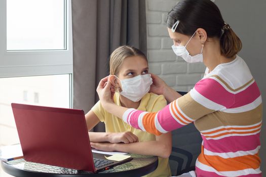 Mom puts on a girl a medical face mask while sitting in front of a computer at home