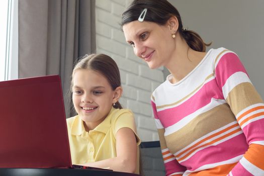 Happy girl and girl are sitting at the table and looking at the laptop screen.