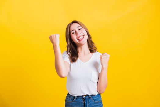 Asian Thai happy portrait beautiful cute young woman standing wear t-shirt makes raised fists up celebrating her winning success looking to camera isolated, studio shot on yellow background with copy space