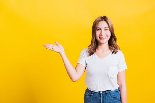 Asian Thai happy portrait beautiful cute young woman standing wear white t-shirt holding something on palm away side looking to camera, studio shot isolated on yellow background with copy space