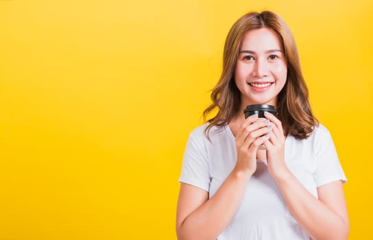 Portrait Asian Thai beautiful happy young woman wear white t-shirt standing smiling holding take away coffee paper cup, studio shot isolated on yellow background, with copy space