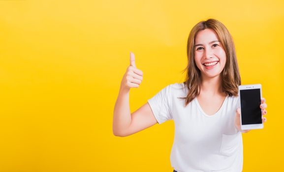 Portrait Asian Thai beautiful happy young woman standing smile, holding blank screen mobile phone and showing thumbs up gesture, shoot photo in studio focus face on yellow background, with copy space