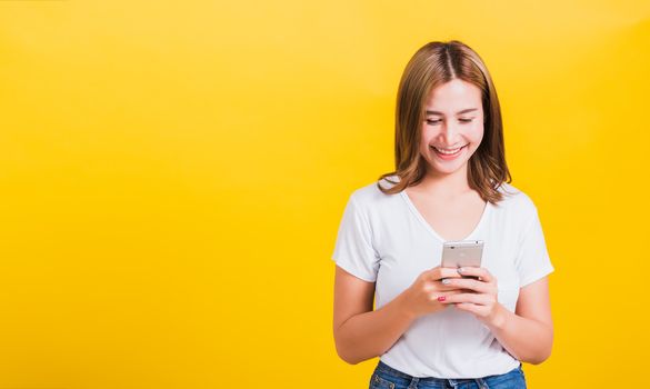 Asian Thai portrait happy beautiful cute young woman smile standing playing game or writing SMS message on smartphone looking to the phone, studio shot isolated on yellow background with copy space