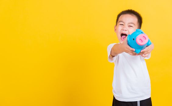 Asian Thai happy portrait cute little cheerful child boy smile holding piggy bank and looking camera, studio shot isolated on yellow background with copy space