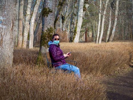 Lady and coronavirus protection. Sad Woman sitting alone on bench in park wearing mask to avoid infectious. Corona virus, or Covid 19, is spreading all over the world. Receives bad news on her phone.