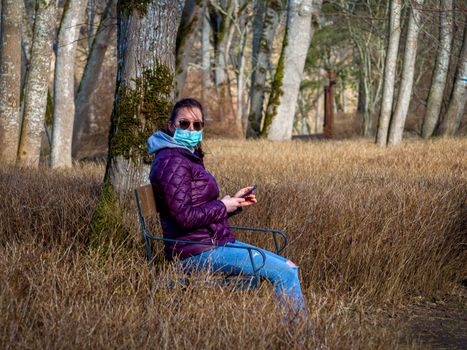 Lady and coronavirus protection. Sad Woman sitting alone on bench in park wearing mask to avoid infectious. Corona virus, or Covid 19, is spreading all over the world. Receives bad news on her phone.