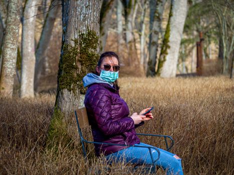 Lady and coronavirus protection. Sad Woman sitting alone on bench in park wearing mask to avoid infectious. Corona virus, or Covid 19, is spreading all over the world. Receives bad news on her phone.