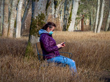 Lady and coronavirus protection. Sad Woman sitting alone on bench in park wearing mask to avoid infectious. Corona virus, or Covid 19, is spreading all over the world. Receives bad news on her phone.