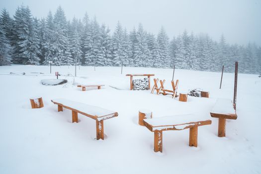 Winter forest camp, woden benches and fireplace covered in snow, snowy landscape.