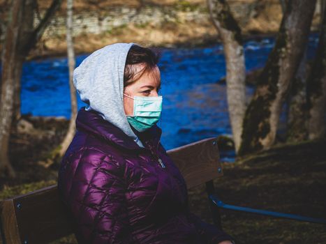 Lady and coronavirus protection. Sad Woman sitting alone on bench in park wearing mask to avoid infectious. Corona virus, or Covid 19, is spreading all over the world. Receives bad news on her phone.