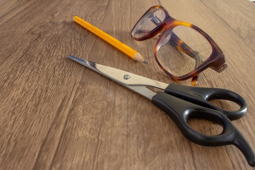 Scissor next to a pencil and a par of glasses on top of a wooden desk