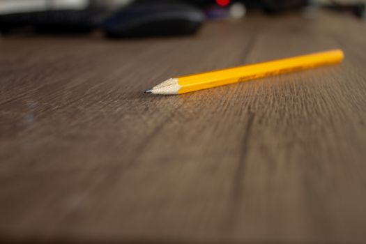 Focus on a yellow pencil on top of a wooden desk, with a computer on the background