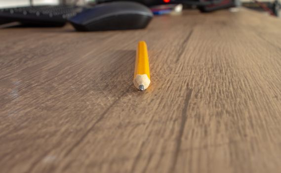 Focus on a yellow pencil on top of a wooden desk, with a computer on the background