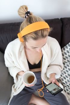 Woman at home relaxing on sofa couch drinking tea from white cup, listening to relaxing music while reading emails and using social media on mobile phone device. Stay at home. Social distancing.