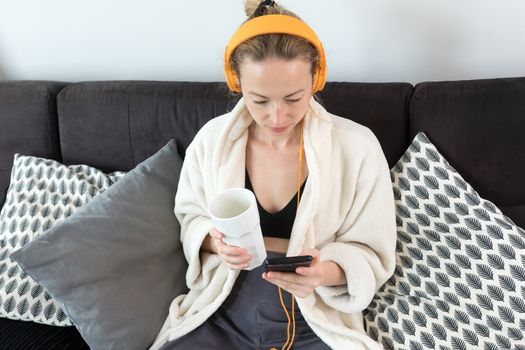 Woman at home relaxing on sofa couch drinking tea from white cup, listening to relaxing music while reading emails and using social media on mobile phone device. Stay at home. Social distancing.