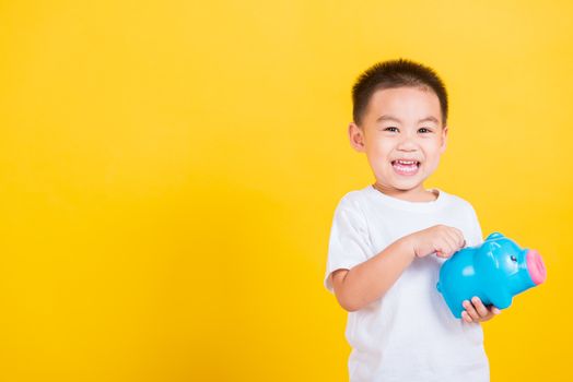 Asian Thai happy portrait cute little cheerful child boy smile putting coin money to the piggy bank and looking camera, studio shot isolated on yellow background with copy space