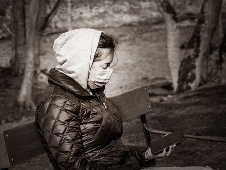 Lady and coronavirus protection. Sad Woman sitting alone on bench in park wearing mask to avoid infectious. Corona virus, or Covid 19, is spreading all over the world. Receives bad news on her phone.