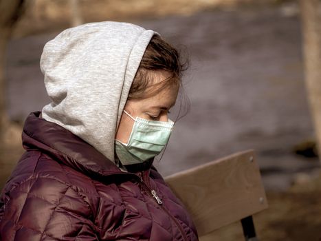 Lady and coronavirus protection. Sad Woman sitting alone on bench in park wearing mask to avoid infectious. Corona virus, or Covid 19, is spreading all over the world. Receives bad news on her phone.