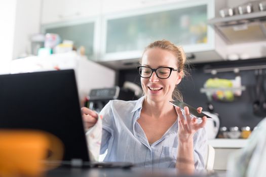 Stay at home and social distancing. Woman in her casual home clothing working remotly from kitchen dining table. Video chatting using social media with friend, family, business clients or partners.