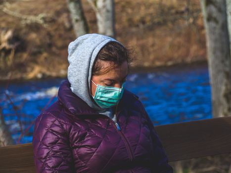 Lady and coronavirus protection. Sad Woman sitting alone on bench in park wearing mask to avoid infectious. Corona virus, or Covid 19, is spreading all over the world. Receives bad news on her phone.