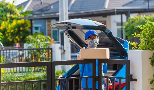 Asian Young delivery man courier online with box in blue cap and t-shirt uniform he protective face mask, service customer in front of the house under curfew quarantine pandemic coronavirus COVID-19