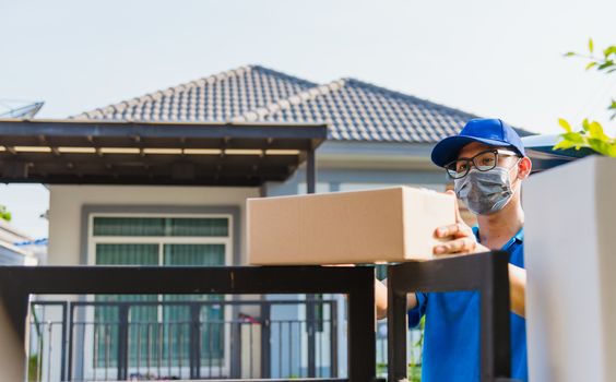 Asian Young delivery man courier online with box in blue cap and t-shirt uniform he protective face mask, service customer in front of the house under curfew quarantine pandemic coronavirus COVID-19