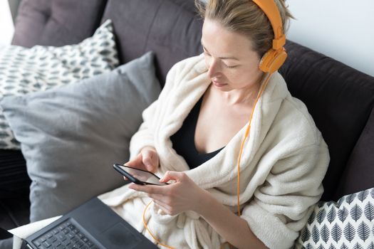 Woman in bathrobe being comfortable at her home sofa, using social media apps on phone for video chatting and stying connected with her loved ones. Social distancing. Stay at home.