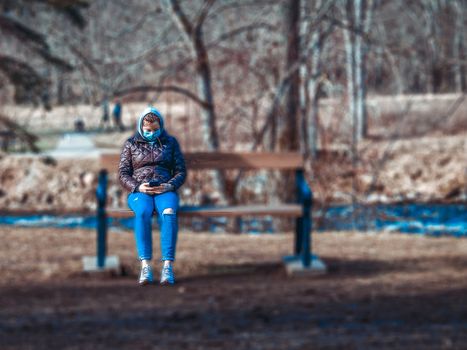 Lady and coronavirus protection. Sad Woman sitting alone on bench in park wearing mask to avoid infectious. Corona virus, or Covid 19, is spreading all over the world. Receives bad news on her phone.