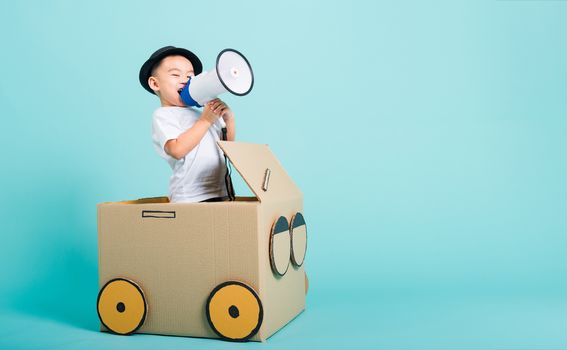 Happy Asian children boy smile in driving play car creative by a cardboard box imagination with megaphone, summer holiday travel concept, studio shot on blue background with copy space for text