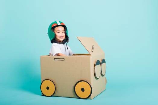 Happy Asian children boy with Helmet smile in driving play car creative by a cardboard box imagination, summer holiday travel concept, studio shot on blue background with copy space for text