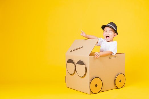 Portrait happy Asian cute little children boy smile so happy wearing white T-shirt driving car creative by cardboard and pointing finger, studio shot on yellow background with copy space