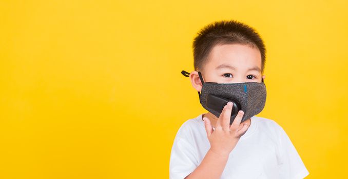 Asian Thai happy portrait cute little cheerful child boy wearing mask protective for covid-19 or pm2.5 dust he looking to camera, studio shot isolated on yellow background with copy space