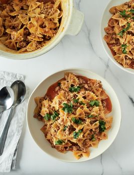 Pasta Farfalle with tomato sauce and ground meat in plate on white marble tabletop. Idea and recipes for easy, simple lunch or dinner one-pot meat goulash. Top down view or flat lay. Vertical