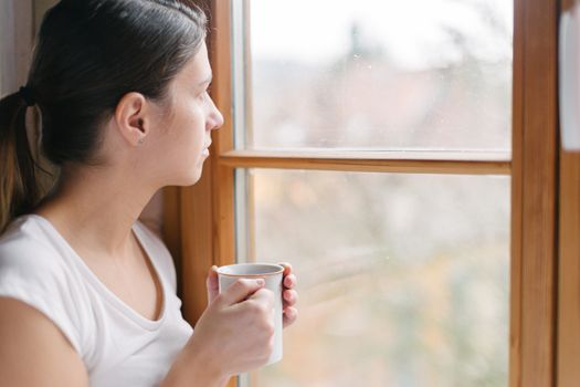 Interior shot of young woman holding a cup of tea looking at the outside world through the window gazing into the distance.