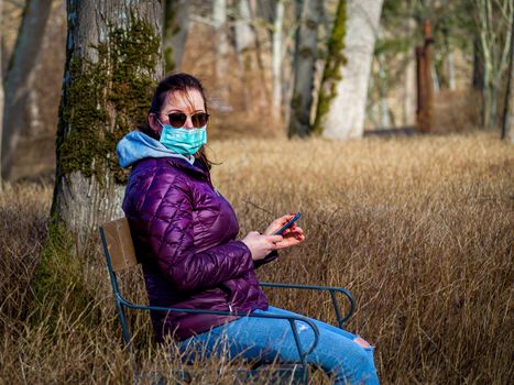 Lady and coronavirus protection. Sad Woman sitting alone on bench in park wearing mask to avoid infectious. Corona virus, or Covid 19, is spreading all over the world. Receives bad news on her phone.