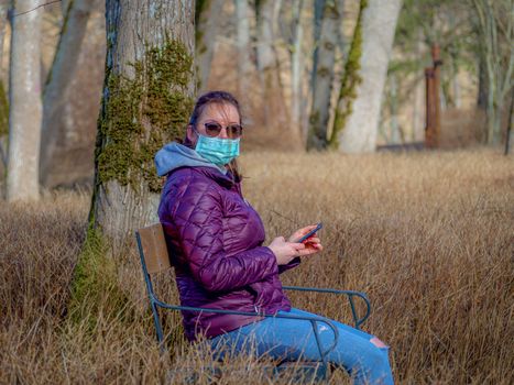 Lady and coronavirus protection. Sad Woman sitting alone on bench in park wearing mask to avoid infectious. Corona virus, or Covid 19, is spreading all over the world. Receives bad news on her phone.