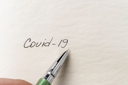 The hand writing Covid-19 on empty sheet of paper. Covid-19 inscription on a blank empty sheet of paper in a notebook. Nearby lies a fountain pen. A lot of space for inscriptions and copyspace. The virus is dangerous. Blank for inscriptions. Template for article or cover.