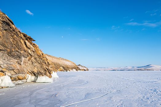 The rocky mountains are covered with snow. The blue color of the sky contrasts with the brown color of mountains and ground. Beautiful scenery of mountains, lake and skies. Lake Baikal, Russia.