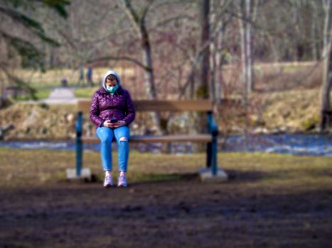 Lady and coronavirus protection. Sad Woman sitting alone on bench in park wearing mask to avoid infectious. Corona virus, or Covid 19, is spreading all over the world. Receives bad news on her phone.