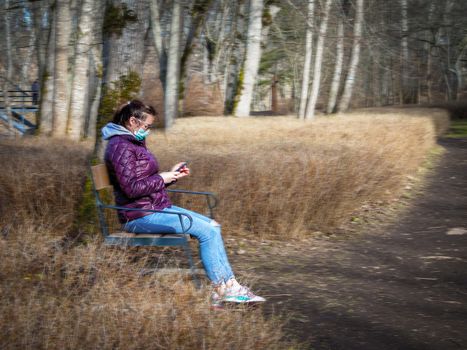 Lady and coronavirus protection. Sad Woman sitting alone on bench in park wearing mask to avoid infectious. Corona virus, or Covid 19, is spreading all over the world. Receives bad news on her phone.