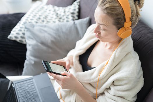 Woman in bathrobe being comfortable at her home sofa, using social media apps on phone for video chatting and stying connected with her loved ones. Social distancing. Stay at home.