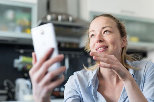 Young smiling cheerful woman indoors at home kitchen using social media apps on phone for video chatting and stying connected with her loved ones. Stay at home, social distancing lifestyle.