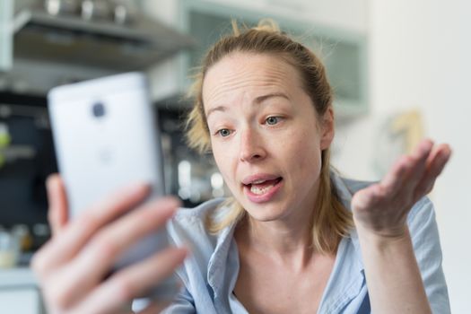 Young smiling cheerful woman indoors at home kitchen using social media apps on phone for video chatting and stying connected with her loved ones. Stay at home, social distancing lifestyle.