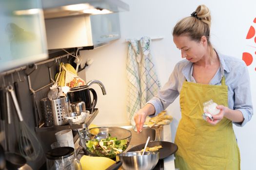 Stay at home housewife woman cooking in kitchen, salting dish in a saucepan, preparing food for family dinner. Blonde caucasian model in her 30is, real cooking not looking in camera.