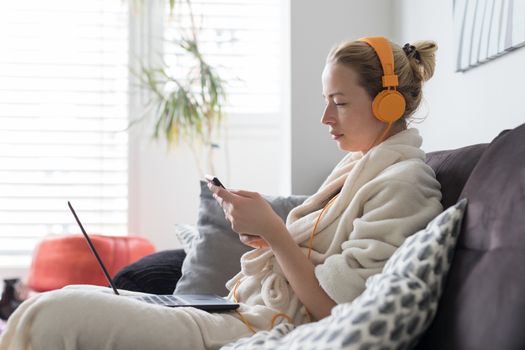 Woman in bathrobe being comfortable at her home sofa, using social media apps on phone for video chatting and stying connected with her loved ones. Social distancing. Stay at home.
