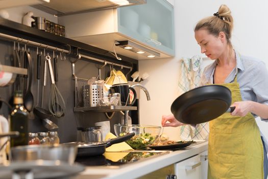 Stay at home housewife woman cooking in kitchen, stir frying dish in a saucepan, preparing food for family dinner. Blonde caucasian model in her 30is, real cooking not looking in camera.