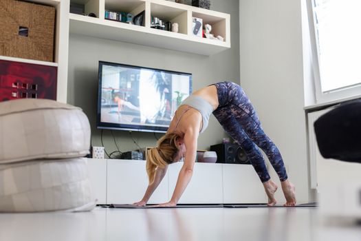 Attractive sporty woman working out at home, doing pilates exercise in front of television in her living room. Social distancing. Stay healthy and stay at home during corona virus pandemic.